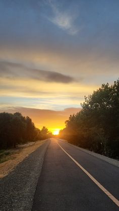 the sun is setting on an empty road with trees in the foreground and clouds in the background