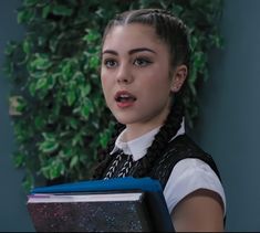 a woman with braids holding a binder in front of a green plant wall