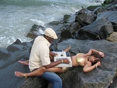 a man sitting next to a woman on top of a rock near the ocean with an open laptop computer