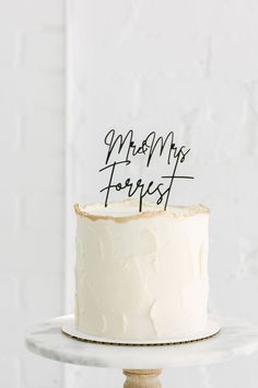 a white cake with black writing on it sitting on top of a wooden stand in front of a white brick wall