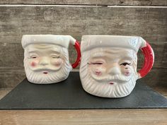 two ceramic santa mugs with red handles on top of a wooden table next to each other