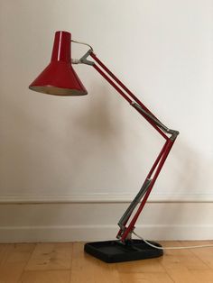 a red desk lamp sitting on top of a hard wood floor next to a white wall