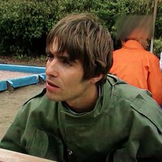 a young man sitting at a table in front of other people and looking off into the distance