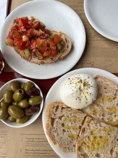 two white plates topped with bread and olives next to an assortment of different types of food