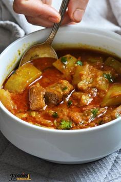 a person holding a spoon over a bowl of food with meat and potatoes in it