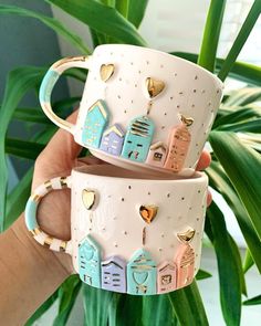 two coffee mugs with houses and hearts on them are held in front of a potted plant