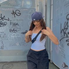 a young woman is standing in front of a graffiti covered wall wearing a baseball cap and suspenders