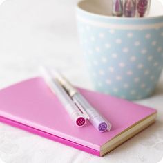 two pens sitting on top of a pink notebook next to a cup