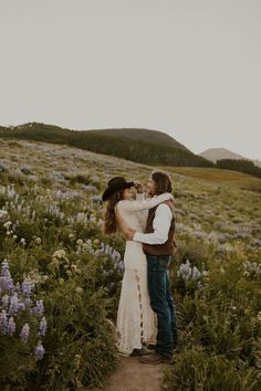 a man and woman standing in the middle of a field with wildflowers at sunset