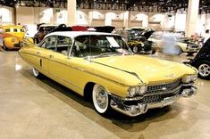 an old yellow car is parked in a showroom with other cars and people looking at it