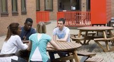 four people are sitting at a table outside in front of a brick building and talking to each other