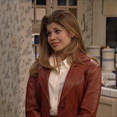 a woman standing in a kitchen wearing a brown jacket and white shirt with her hands on her hips