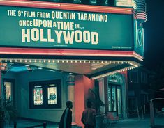 two people standing in front of a movie theater at night with the hollywood sign lit up