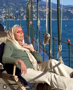 an old woman sitting on a chair next to the ocean with red sunglasses and a blanket over her head