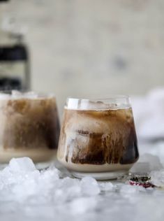 two glasses filled with iced coffee sitting on top of snow covered ground next to a bottle