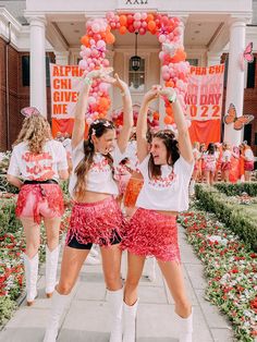 two girls in pink and white cheerleader outfits hold up balloons above their heads as they walk down a sidewalk