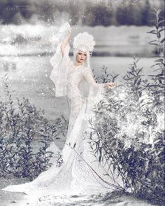 a woman in a white dress is standing by some bushes and trees with her arms outstretched