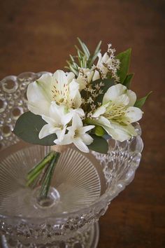 white flowers are in a glass vase on a clear plate, with greenery and water droplets