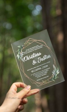 a person holding up a wedding card in front of the camera with greenery on it