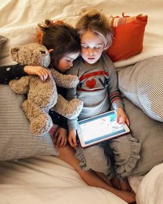 two children are sitting on a bed and one is holding a tablet while the other holds a teddy bear