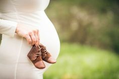 a pregnant woman wearing brown shoes and holding her belly with both hands while standing in the grass