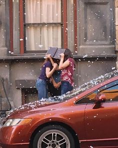 two people standing on the roof of a car in front of a building with snow falling all over it