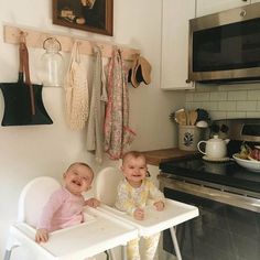 two babies sitting in high chairs next to an oven and microwave with utensils hanging on the wall