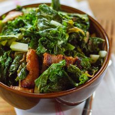 a brown bowl filled with greens and meat on top of a wooden table next to a fork