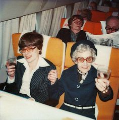 two people sitting on an airplane holding drinks and reading the paper while another person holds up a newspaper