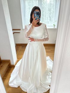 a woman in a white dress taking a selfie with her cell phone while wearing a long sleeved wedding gown