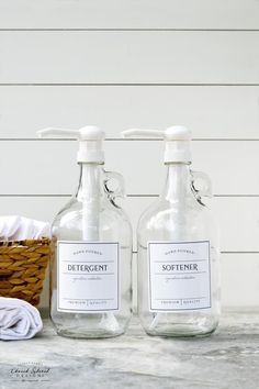 two clear bottles with white labels on them sitting next to a basket and towel dispenser