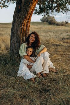 two women are sitting under a tree in the grass and one woman is holding her child