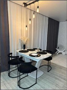 a white table with four black chairs in front of a curtained wall and two lights hanging from the ceiling