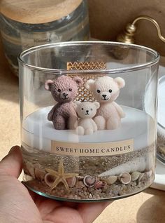 three teddy bears in a glass jar with sand and shells around it that says sweet home candles