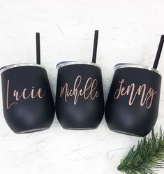 three black tumblers with pink lettering on them sitting next to a pine branch and evergreen