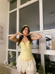 a woman standing in front of a window wearing a yellow shirt and white ruffled skirt