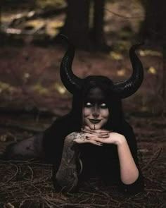 a woman with black makeup and horns on her face is laying in the woods while holding her hands to her chest