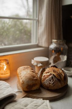 two loaves of bread sit on a cutting board next to a candle and window