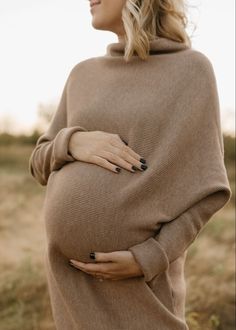 a pregnant woman wearing a brown sweater and black nail polish stands in a field with her hands on her stomach