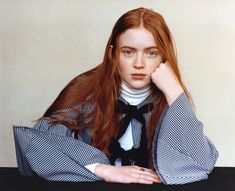 a woman with long red hair wearing a black and white checkered shirt sitting at a table