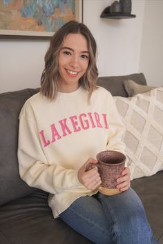 a woman sitting on a couch holding a coffee cup and smiling at the camera while wearing a sweater that says lakegirl