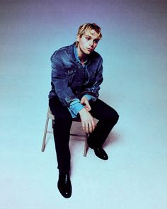 a man sitting on top of a chair wearing a denim shirt and black pants with his legs crossed