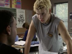 a young man sitting at a desk in front of another person who is holding papers