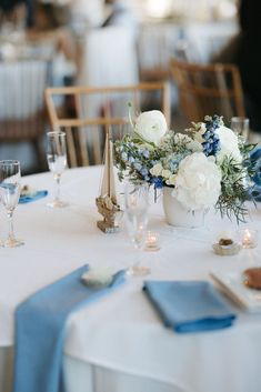 Image of white and blue coastal wedding centerpiece floral designs featuring peonies and ranunculus. The image is a close up of a table at the reception for a wedding at the Coonamessett. The table also has pale blue napkins and sailboat table numbers. Blue Floral Centerpieces Simple, Coastal Chic Wedding Flowers, Blue And White Low Centerpiece, Coastal Chic Wedding Tables Round, Blue And White Centerpieces, White Peonies Centerpiece, Small Blue And White Floral Centerpieces, Coastal Tablescapes, Coastal Wedding Centerpieces