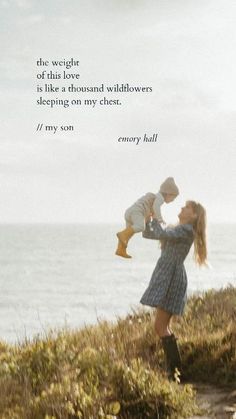 a woman holding a baby on top of a grass covered hillside next to the ocean