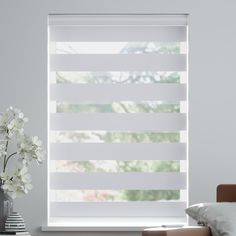 a window with white blinds and flowers in front of it on a table next to a couch