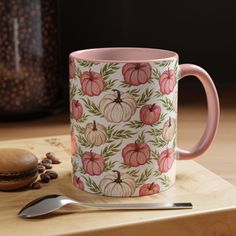 a pink coffee mug sitting on top of a wooden table next to a spoon and cup