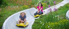 two children riding on the back of a water slide in a flowered garden with their mother