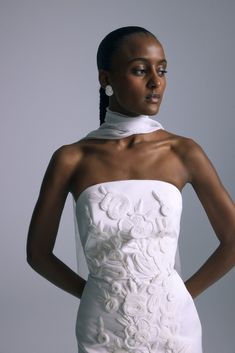 a woman wearing a white dress with flowers on the back and neckline, standing in front of a gray background