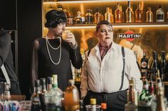 two men standing next to each other at a bar with bottles on the wall behind them